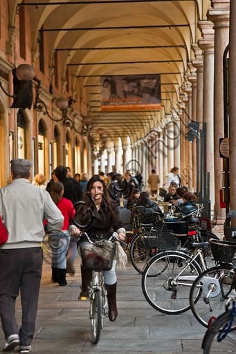Veduta dei portici storici in Via Emilia Centro, a Modena, con persone che passeggiano e circolano in bicicletta sotto le arcate.