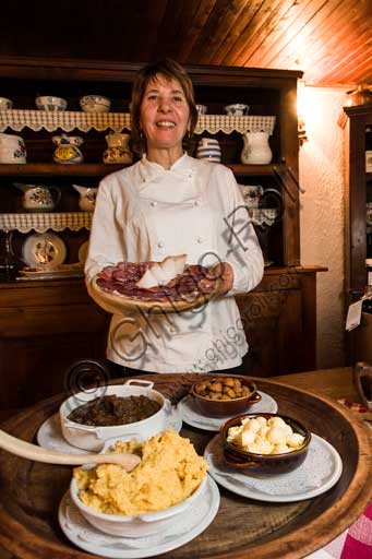   Gressoney La Trinité, Restaurant "La Capanna di Carla": Susanna, cook and owner, shows a selection of cold cuts. On the table, some typical dishes such as polenta, venison stew and chestnuts.
