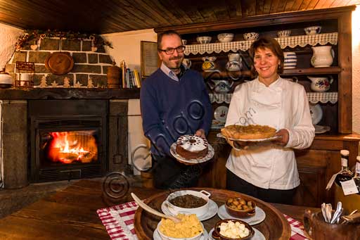   Gressoney La Trinité, Restaurant "La Capanna di Carla". Susanna and Gigi, the owners, show two cakes. On the table, some typical dishes such as polenta, venison stew and chestnuts.