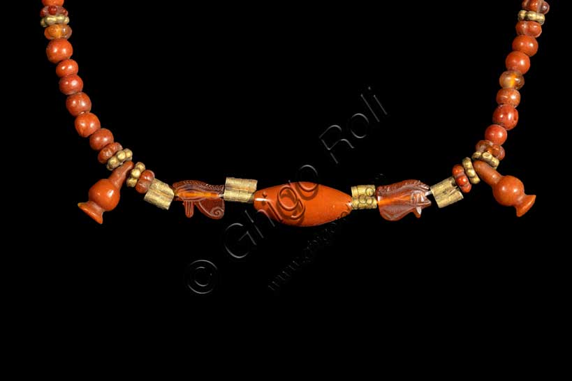 Museo Civico Archeologico di Bologna: collana con melagrane.  Nuovo Regno: XVIII-XX dinastia (1539-1075 a.C.).Inventario EG 3103 Particolare.La collana, costituita da vaghi configurati a melagrana e a rosetta, da perline sferiche e tubicini in corniola, ai quali si alternano tubicini e rosette in oro, ha al centro un vago ovoidale tra due occhi udjat del dio Horo affrontati, simboli di integrità. Al suo frutto, ricco di chicchi succosi, gli Egiziani associano da subito l'idea della fertilità e della rinascita, e perciò indossare una collana come questa era propiziatorio di una lunga vita, terrena o ultraterrena. Anche la scelta della corniola, simile nel colore alle tonalità arancio-rossastre della melagrana, ne rafforzava la valenza magica.