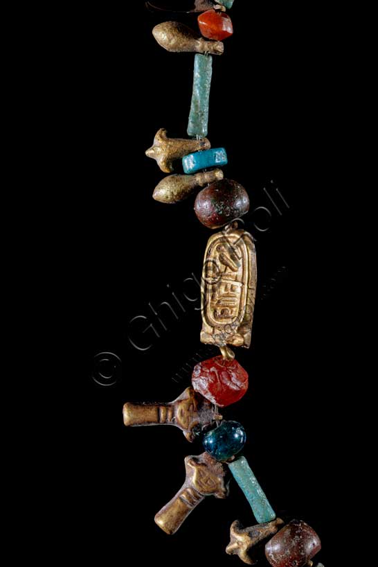 Museo Civico Archeologico di Bologna: collana con cartiglio del dio Aton, dettaglio del cartiglio.Inventario EG 3069.Nuovo Regno: XVIII dinastia, regno di Akhenaton (1353 - 1336 a.C.).Fra i vari elementi che compongono questa collana -perline alternate a tubetti, testine della dea Hathor ed elementi vegetali- spicca un piccolo cartiglio in lamina d’oro su cui è scritto a sbalzo uno dei nomi del dio Aton. Questa divinità, che rappresenta per gli Egiziani il disco del sole, assunse grande importanza durante il regno del faraone Amenhotep IV/Akhenaton (1353-1336 a.C.), soppiantando per un breve periodo il culto dell’importantissimo dio tebano Amon. Particolare.