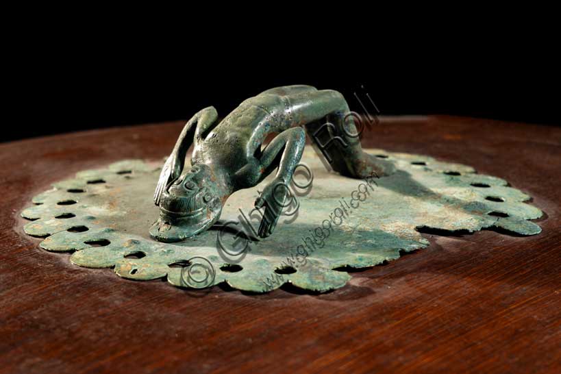 Museo Civico Archeologico di Bologna: corredo funerario dalla tomba 103 della necropoli De Luca.Al centro è la cista in bronzo inventario 16537. La cista era originariamente costituita da un corpo di legno rivestito di cuoio, non conservato in originale ma ricostruito, sul quale era montata una lamina traforata di bronzo.Il contenitore è decorato con elementi di bronzo: i piedi sono a forma di zampe di leone, a metà dell'altezza del corpo sono inserite delle piccole teste di ariete e sul coperchio la presa è costituita da una piccola e graziosa figura di atleta che esegue il ponte.Nella cista la donna etrusca riponeva i propri oggetti da toilette: l'alàbastron, cioè il contenitore per olii profumati, il pettine in osso e lo specchio di bronzo, le pinzette e gli altri materiali per la cura del corpo.Metà del V secolo a.C. Inv. 16537.Particolare del coperchio con la presa  costituita da una piccola e graziosa figura di atleta che esegue il ponte.