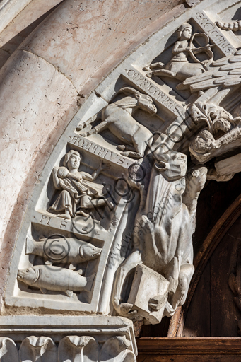 Foligno, Cattedrale di San Feliciano, la facciata: il portale. Particolare del fregio. Il portale centrale, opera dei maestri Rodolfo e Binello, mostra un disco solare nella lunetta, in cui si trova l'iscrizione con la data 1201, anno del completamento della facciata; nella facciata interna degli stipiti si trovano poi i rilievi con l'Imperatore Ottone IV di Brunswick e papa Innocenzo III, mentre il giro interno dell'arco è decorato dai Simboli degli evangelisti e i Segni dello zodiaco; nel giro esterno una fascia di mosaici cosmateschi. Il portale ligneo intagliato è del 1620. 