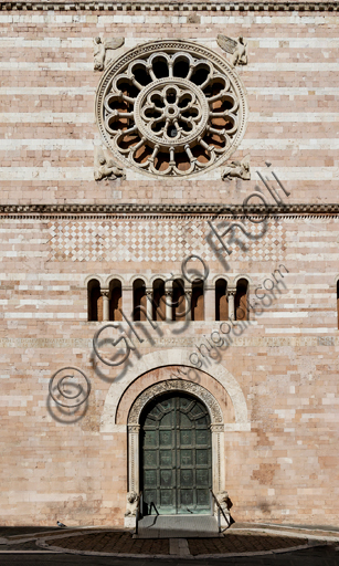 Foligno, Cattedrale di San Feliciano, la facciata: particolare del portale e del rosone romanico. Il portale centrale, opera dei maestri Rodolfo e Binello, mostra un disco solare nella lunetta, in cui si trova l'iscrizione con la data 1201, anno del completamento della facciata; nella facciata interna degli stipiti si trovano poi i rilievi con l'Imperatore Ottone IV di Brunswick e papa Innocenzo III, mentre il giro interno dell'arco è decorato dai Simboli degli evangelisti e i Segni dello zodiaco; nel giro esterno una fascia di mosaici cosmateschi. Il portale ligneo intagliato è del 1620. 