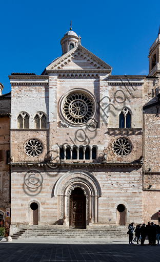 Foligno, Cattedrale di San Feliciano: la facciata con i tre rosoni e il portale. Il portale centrale, opera dei maestri Rodolfo e Binello, mostra un disco solare nella lunetta, in cui si trova l'iscrizione con la data 1201, anno del completamento della facciata; nella facciata interna degli stipiti si trovano poi i rilievi con l'Imperatore Ottone IV di Brunswick e papa Innocenzo III, mentre il giro interno dell'arco è decorato dai Simboli degli evangelisti e i Segni dello zodiaco; nel giro esterno una fascia di mosaici cosmateschi. Il portale ligneo intagliato è del 1620. 