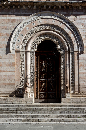 Foligno, Cattedrale di San Feliciano, la facciata: il portale. Il portale centrale, opera dei maestri Rodolfo e Binello, mostra un disco solare nella lunetta, in cui si trova l'iscrizione con la data 1201, anno del completamento della facciata; nella facciata interna degli stipiti si trovano poi i rilievi con l'Imperatore Ottone IV di Brunswick e papa Innocenzo III, mentre il giro interno dell'arco è decorato dai Simboli degli evangelisti e i Segni dello zodiaco; nel giro esterno una fascia di mosaici cosmateschi. Il portale ligneo intagliato è del 1620. 