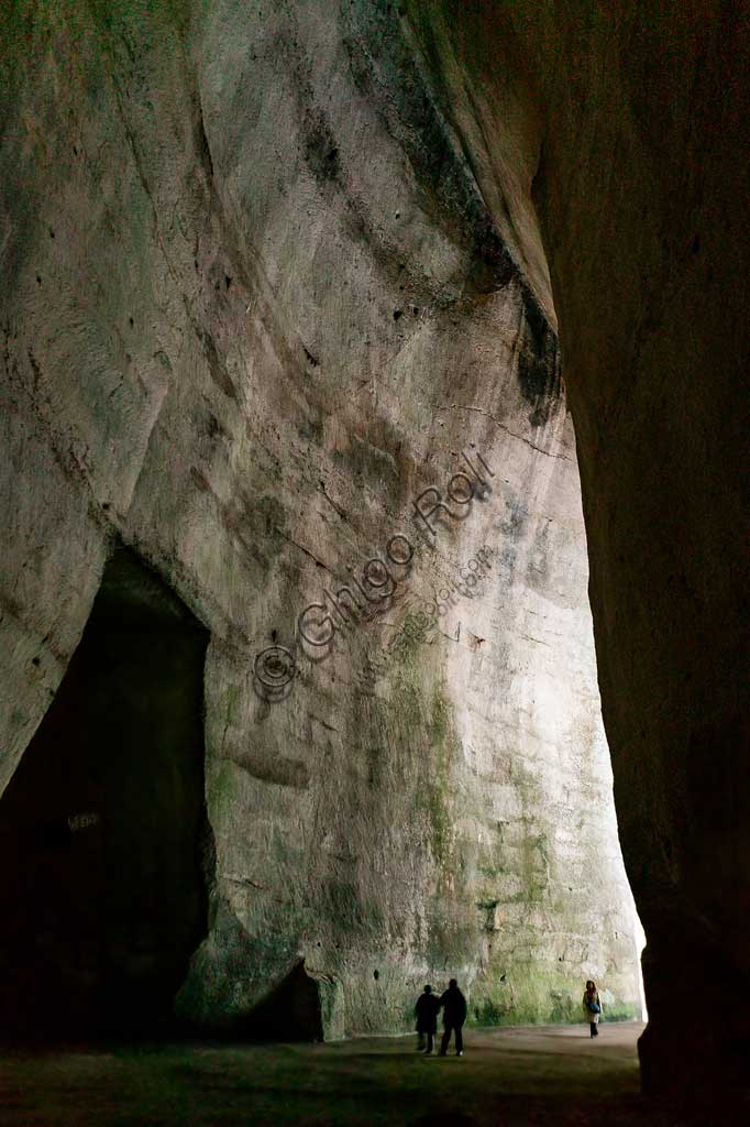 Siracusa, Parco archeologico della Neapolis di Siracusa:  l'ingresso dell'Orecchio di Dionigi (o Dionisio).L'Orecchio di Dionisio (o Orecchio di Dionigi) è una grotta artificiale che si trova nell'antica cava di pietra detta latomia del Paradiso, sotto il Teatro Greco di Siracusa. Scavata nel calcare, è alta circa 23 m. e larga dai 5 agli 11 m., e si sviluppa in profondità per 65 m., con un andamento ad S che lo rende anche un luogo di amplificazione acustica dei suoni (sino a 16 volte).Secondo la leggenda, la sua particolare forma ad orecchio d'asino fece coniare al pittore Caravaggio l'espressione Orecchio di Dionisio. Secondo la tradizione infatti il tiranno Dionisio fece scavare la grotta dove rinchiudeva i prigionieri, e appostandosi all'interno di una cavità superiore ascoltava i loro discorsi. 