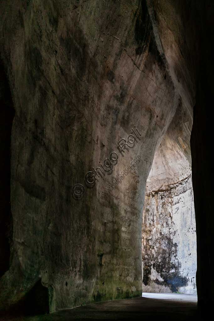 Syracuse, The Archaeological Park of the Neapolis of Syracuse: the entrance to the Ear of Dionysius (or Dionysus).The Ear of Dionysius (or Ear of Dionysius) is an artificial cave located in the ancient stone quarry called the Latomia of Paradise, under the Greek Theatre of Syracuse. Excavated in limestone, it is about 23 m high. and 5 to 11 m. wide, and 65 m. deep, with an S-shape that makes it a place of acoustic amplification of sounds (up to 16 times).According to the legend, Caravaggio gave it its name. According to tradition, the tyrant Dionysius had the cave excavated and he locked up the prisoners and he listened to their speeches.