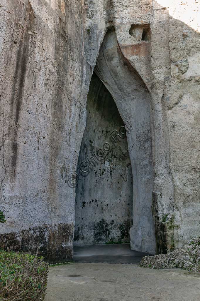 Siracusa, Parco archeologico della Neapolis di Siracusa:  l'ingresso dell'Orecchio di Dionigi (o Dionisio).L'Orecchio di Dionisio (o Orecchio di Dionigi) è una grotta artificiale che si trova nell'antica cava di pietra detta latomia del Paradiso, sotto il Teatro Greco di Siracusa. Scavata nel calcare, è alta circa 23 m. e larga dai 5 agli 11 m., e si sviluppa in profondità per 65 m., con un andamento ad S che lo rende anche un luogo di amplificazione acustica dei suoni (sino a 16 volte).Secondo la leggenda, la sua particolare forma ad orecchio d'asino fece coniare al pittore Caravaggio l'espressione Orecchio di Dionisio. Secondo la tradizione infatti il tiranno Dionisio fece scavare la grotta dove rinchiudeva i prigionieri, e appostandosi all'interno di una cavità superiore ascoltava i loro discorsi. 