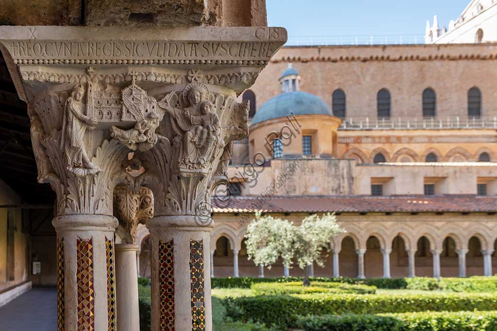 Monreale, Duomo, chiostro del monastero benedettino (XII secolo):    il capitello W 8, faccia Sud;  "L'imperatore Guglielmo II offre la Cattedrale alla Vergine col Bambino". Sullo sfondo, il fianco meridionale della Cattedrale e le aiuole con un ulivo.