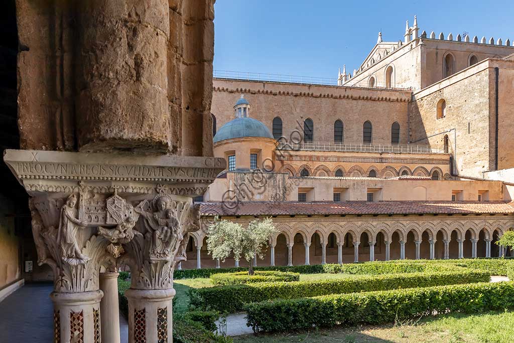 Monreale, Duomo, chiostro del monastero benedettino (XII secolo):    il capitello W 8, faccia Sud;  "L'imperatore Guglielmo II offre la Cattedrale alla Vergine col Bambino". Sullo sfondo, il fianco meridionale della Cattedrale e le aiuole con un ulivo.