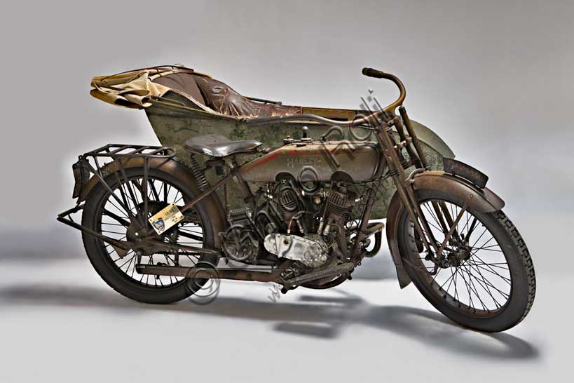 Moto d'epoca “Harley-Davidson T - Side”Marca: Harley-Davidsonmodello: T Sidenazione: U.S.A. - Milwaukeeanno: 1917condizioni: conservatacilindrata: 1000 (alesaggio e corsa 84,1 x 88,9)motore: bicilindrico a V di 45°cambio: a tre rapportiFondata nel 1903 la H-D ha accompagnato tutta la storia degli Stati Uniti fino ad oggi, dalle prime moto militari usate nelle schermaglie di confine contro Pancho Villa, alle grandi forniture per l'esercito che nel 1917 era entrato in guerra sul fronte europeo, dai mezzi da lavoro (i "tricicli" dei lattai e dei postini americani) alle grandi bicilindriche della polizia. Già nel '20 era il primo produttore di motociclette al mondo.Questo elegante sidecar del 1917, integralmente conservato e perfettamente funzionante, mostra già il bicilindrico a V che caratterizza ancor oggi la produzione della Harley. Ha distribuzione a valvole contrapposte (scarico laterale, aspirazione in testa).