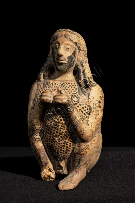 Museo Civico Archeologico di Bologna: Balsamario corinzio configurato.Prediletti della produzione corinzia sono i piccoli contenitori, aryballoi e alabastra e a partire dall’inizio del VI sec. a.C. un tipo di anfora miniaturizzata, l’amphoriskos, tutti destinati a contenere profumi e unguenti. Nell’ambito delle forme destinate alla cosmesi una tipologia del tutto peculiare è quella degli unguentari plastici, per lo più a forma di animale e destinati soprattutto all’esportazione. A questo stesso scopo era destinato anche il piccolo unguentario a forma di uomo accovacciato, con barba e lunghi capelli ricadenti sulla spalle e sulla schiena. Indossa una veste decorata a fitti puntini che probabilmente riproduce una pelle ferina e presenta una coppia di fori a fianco del collo destinati alla sospensione del recipiente. Inv. G 599.