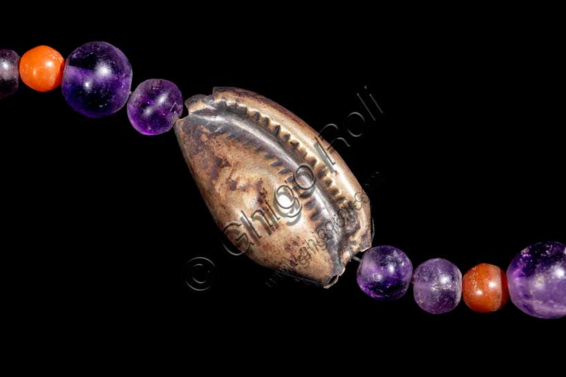 Museo Civico Archeologico di Bologna: collana con cipree, ametista, corniola e argento.Inventario EG 3077Ornamento personale – Medio Regno: XII dinastia (1938-1759 a.C.) Lo splendido gioiello è composto da una serie di perline in corniola e in ametista, alternate a conchiglie cipree in argento lavorate a sbalzo, che ne rappresentano l’elemento caratterizzante. Grazie al confronto con esemplari analoghi rinvenuti in contesti funerari a el-Lisht, Dahshur e Tebe ovest, il monile è databile con sicurezza alla XII dinastia (1938-1759 a.C.). Considerato dalla stragrande maggioranza degli studiosi una collana, questo raro gioiello non è escluso potesse essere usato quale cintura da indossare sui fianchi, non solo per esaltare l’armoniosa nudità di un giovane corpo femminile, ma anche per propiziarne una sicura fertilità, evocata dalle conchiglie cipree che ne sono un simbolo tradizionale. Particolare.