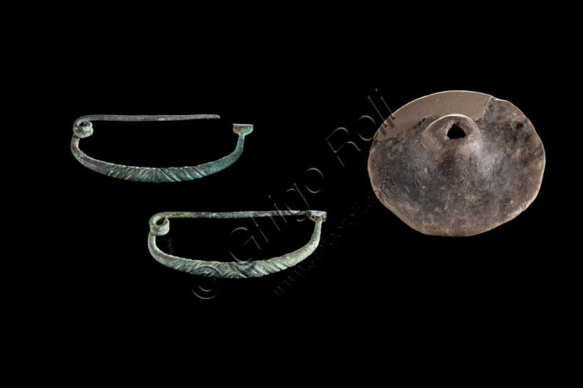 Museo Civico Archeologico di Bologna: coppia di fibule ad arco ribassato ingrossato in bronzo e  piccola ruota in terracotta;  Bronzo Finale Ferro 1. inv. SBAER 10890.