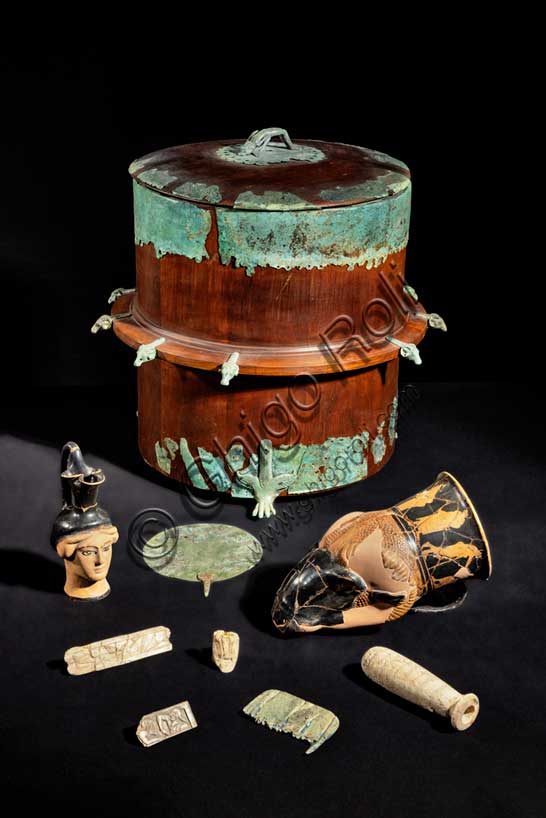 Museo Civico Archeologico di Bologna: corredo funerario dalla tomba 103 della necropoli De Luca.Al centro è la cista in bronzo inventario 16537. La cista era originariamente costituita da un corpo di legno rivestito di cuoio, non conservato in originale ma ricostruito, sul quale era montata una lamina traforata di bronzo.Il contenitore è decorato con elementi di bronzo: i piedi sono a forma di zampe di leone, a metà dell'altezza del corpo sono inserite delle piccole teste di ariete e sul coperchio la presa è costituita da una piccola e graziosa figura di atleta che esegue il ponte.Nella cista la donna etrusca riponeva i propri oggetti da toilette: l'alàbastron, cioè il contenitore per olii profumati, il pettine in osso e lo specchio di bronzo, le pinzette e gli altri materiali per la cura del corpo.Metà del V secolo a.C. Inv. 16537.