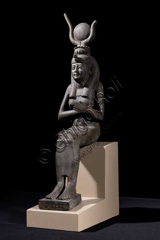 Museo Civico Archeologico di Bologna: Isi (Iside) seduta in trono.Statuetta di Epoca Tarda: XXVI-XXX dinastia (664-342 a.C.). Bronzo con agemina in oro.Inventario EG 248/9.Fra tutte le divinità femminili del pantheon egiziano Isi è la dea per eccellenza, la più conosciuta, “la grande di magia”, soprattutto durante l’Antico Regno (2705-2225 a.C.), quando il suo nome e la sua immagine compaiono raramente sugli edifici religiosi e nei luoghi di culto domestico. Introdotta con un ruolo da protagonista nella leggenda di Osiri, forse attorno al 2500 a.C., le caratteristiche di grande maga si affiancano ad altre più umane, che permettono al suo culto di sopravvivere all'Egitto faraonico, diffondendosi dal VI secolo a.C. in tutto il bacino del Mediterraneo. Isi si identifica così con la fedele compagna del dio Osiri, esperta nell'arte di assecondarlo, di preparane le esequie, di perpetuarne il culto dopo la morte, così come con il prototipo ideale della donna egiziana che veglia sul proprio figlio Horo, erede al trono di Osiri. La dea, raffigurata in questo bronzetto nel suo ruolo di madre premurosa, siede in trono e porge il seno al figlio Horo, ora scomparso. Alla semplicità della veste, una tunica aderente che esalta le linee sinuose del corpo femminile, si contrappongono la ricchezza degli occhi in agemina, la ieraticità della corona hathorica dal tipico disco solare tra corna bovine a lira e il volume dell’acconciatura a tre bande di capelli, sormontata dalla spoglia di avvoltoio. 