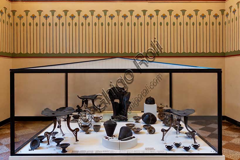 Museo Civico Archeologico di Bologna, sala III: l’allestimento del corredo della tomba 85 dalla necropoli Lippi di Verucchio, uno dei centri Etruschi di maggiore importanza della regione.Tra i materiali di corredo (deposti assieme alle ceneri di un ricco aristocratico) spicca l'imponente trono in legno, decorato ad intaglio, databile al VII secolo a.C., con poggiapiedi. E' il segno più evidente che il defunto avesse rango altissimo.Oltre al trono si segnalano, sempre appartenenti allo stesso corredo, i tre tavolini che ospitavano il vasellame in ceramica contenente le offerte di cibo per il defunto. Tra i cibi è ancora possibile riconoscere uva, nocciole, carne e pesce. A lato del trono vi è un grande elmo in bronzo e vimini, mentre una fibula in bronzo rivestita di preziosa ambra apparteneva all'ornanento personale del defunto.