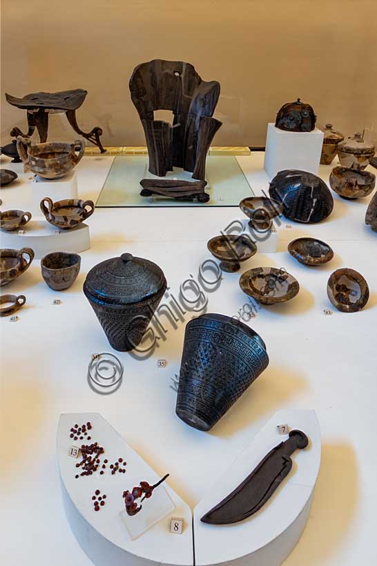 Museo Civico Archeologico di Bologna, sala III: l’allestimento del corredo della tomba 85 dalla necropoli Lippi di Verucchio, uno dei centri Etruschi di maggiore importanza della regione.Tra i materiali di corredo (deposti assieme alle ceneri di un ricco aristocratico) spicca l'imponente trono in legno, decorato ad intaglio, databile al VII secolo a.C., con poggiapiedi. E' il segno più evidente che il defunto avesse rango altissimo.Oltre al trono si segnalano, sempre appartenenti allo stesso corredo, i tre tavolini che ospitavano il vasellame in ceramica contenente le offerte di cibo per il defunto. Tra i cibi è ancora possibile riconoscere uva, nocciole, carne e pesce. A lato del trono vi è un grande elmo in bronzo e vimini, mentre una fibula in bronzo rivestita di preziosa ambra apparteneva all'ornanento personale del defunto.