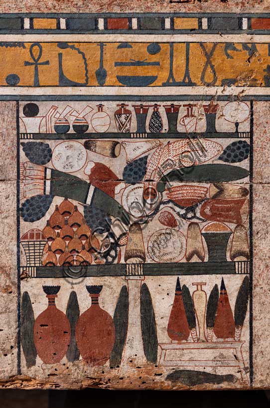 Museo Civico Archeologico di Bologna: sarcofago a cassa a nome di Ibi. Particolare di una serie di pannelli a "falsa porta", motivo ripreso dall'architettura funeraria dell'Antico Regno, che decora questo sarcofago appartenuto alla “signora della casa” Ibi. Medio Regno: XII-XIII dinastia (1938-1640 a.C.); Inv. MCA-EGI-EG_1959. Particolare del pannello centrale caratterizzato da una ricca tavola imbandita. Sotto una fila di piccoli vasetti porta unguenti destinati alla cura del corpo della defunta, sono chiaramente distinguibili pani conici, triangolari e tondi, grappoli d’uva, tranci vari di carne bovina tra cui spiccano la zampa anteriore destra, il cuore e la testa dell’animale, un mazzetto di porri, un mucchietto di fichi, una grossa zucca e due vasi dal contenuto incerto. Ai lati del piede centrale che sorregge la tavola, sono addossati rigogliosi cespi di lattuga in alternanza ad anfore vinarie e olearie. La ricchezza di questa tavola d’offerta funeraria, come sempre, è garantita per l’eternità dall’iscrizione magico-religiosa in caratteri geroglifici che scorre poco sopra, con andamento da destra verso sinistra, e che recita: “Un’offerta funeraria che il sovrano fa al dio Osiri, signore della città di Gedwt (Busiris), grande dio signore della città di Abido perché egli dia un’offerta funeraria consistente in pani, birra, buoi, volatili, tessuti di lino e ogni cosa buona pura e viva per l’anima Ka della devota signora della casa Ibi…”.