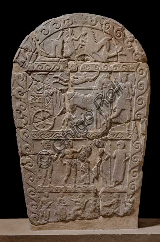 Museo Civico Archeologico di Bologna: Stele funeraria etrusca decorata su quattro registri. Questa stele di dimensioni monumentali, è decorata a bassorilievo su entrambi i lati.ll tema del passaggio del defunto dalla vita alla morte è descritto mediante due scene diverse, in cui compare sempre un demone alato, che, su un lato, “rapisce” il defunto portandolo per mano nell’Aldilà, sull’altro lo guida verso l'oltretomba su una biga trainata da cavalli alati.Questa seconda raffigurazione si inserisce in una delle tre fasce in cui è suddiviso uno dei lati della stele; nelle altre due fasce vi sono, in basso, una rappresentazione di giochi funebri, con un cavaliere etrusco armato che affronta un guerriero nudo con spada e scudo; in alto la lotta tra un serpente ed un ippocampo, animali fantastici che alludono al mondo ultraterreno.Provenienza: Bologna, necropoli della Certosa, tomba 89.Numero di inventario: Ducati 169