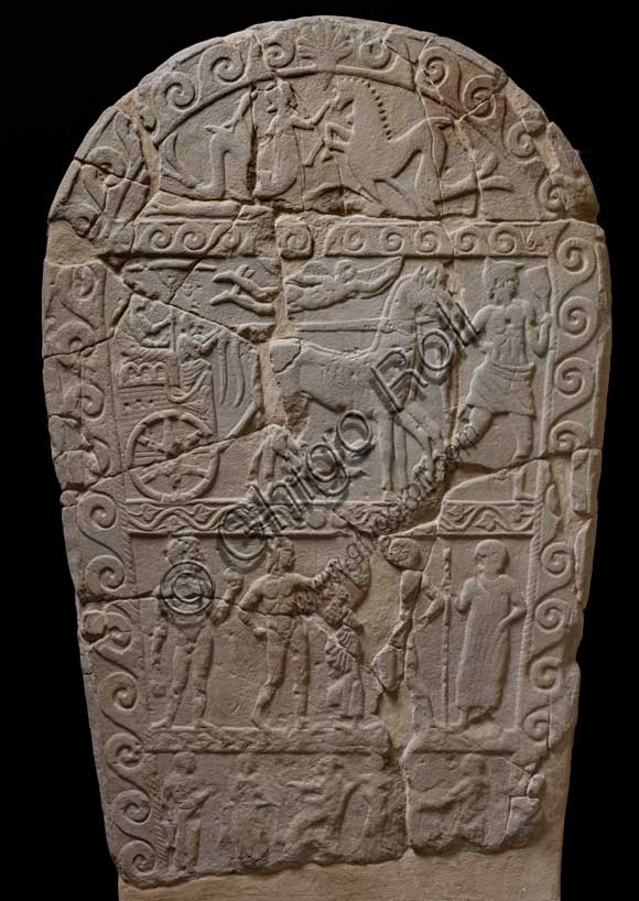Museo Civico Archeologico di Bologna: Stele funeraria etrusca decorata su quattro registri. Questa stele di dimensioni monumentali, è decorata a bassorilievo su entrambi i lati.ll tema del passaggio del defunto dalla vita alla morte è descritto mediante due scene diverse, in cui compare sempre un demone alato, che, su un lato, “rapisce” il defunto portandolo per mano nell’Aldilà, sull’altro lo guida verso l'oltretomba su una biga trainata da cavalli alati.Questa seconda raffigurazione si inserisce in una delle tre fasce in cui è suddiviso uno dei lati della stele; nelle altre due fasce vi sono, in basso, una rappresentazione di giochi funebri, con un cavaliere etrusco armato che affronta un guerriero nudo con spada e scudo; in alto la lotta tra un serpente ed un ippocampo, animali fantastici che alludono al mondo ultraterreno.Provenienza: Bologna, necropoli della Certosa, tomba 89.Numero di inventario: Ducati 169