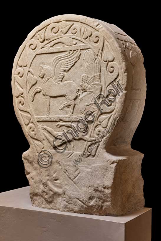 Museo Civico Archeologico di Bologna: stele funeraria etrusca decorata su entrambi i lati e nello spessore, lato A. Proveniente dal sepolcreto dei Giardini Margherita.inv. DUCATI_012Una donna dalla stirpe eroica. La stele Ducati 12, o di Rakvi Satlnei, è stata ritrovata presso la necropoli dei Giardini Margherita, collocata a ovest della città antica, ed è datata attorno al 400 a.C.  Questo lato lato è occupato dal tema del passaggio di status, cioè la morte, reso attraverso l’espediente del viaggio su carro della defunta. Sotto è raffigurato un personaggio maschile nudo che si getta su una spada, riconosciuto come l’eroe omerico Aiace Telamonio. Sulla stele è presente un’iscrizione che, in alcuni punti, è di difficile lettura. Si riesce a distinguere il prenome, Rakvi, e il gentilizio, Satlnei, della donna. È importante sottolineare che si tratterebbe dell’unico caso attestato in tutta Bologna. La seconda parte è molto dibattuta dalla critica ma sembra collegarsi alla raffigurazione di Aiace Telamonio. Infatti, le lettere leggibili sembrano comporre le parole aivas telmuns. Attraverso le informazioni grammaticali in nostro possesso, si è pensato che venga evocato un rapporto genealogico della defunta con l’eroe.