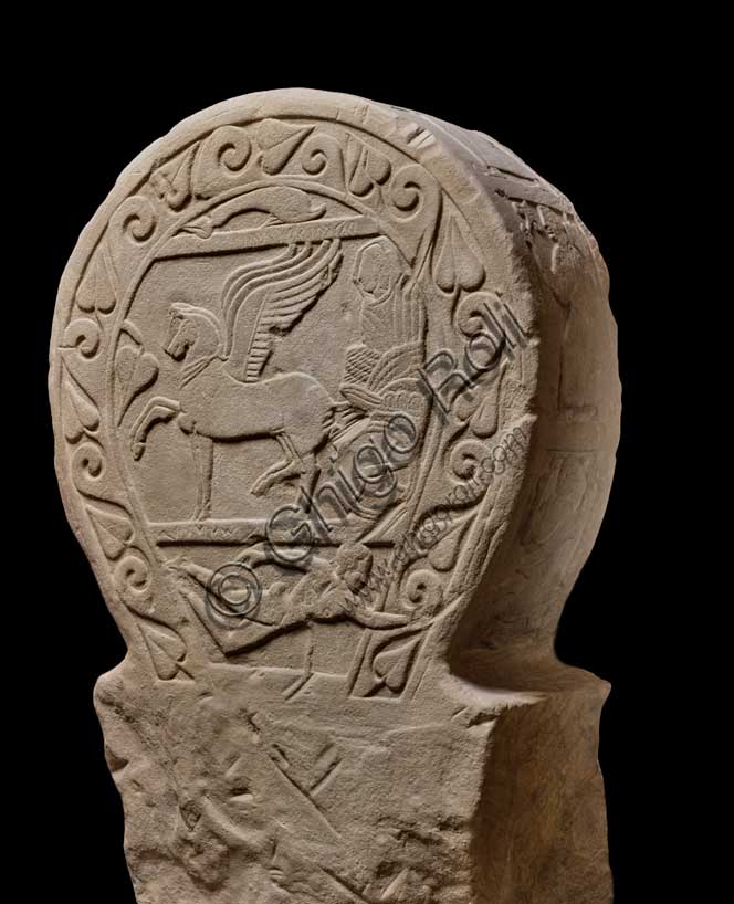 Museo Civico Archeologico di Bologna: stele funeraria etrusca decorata su entrambi i lati e nello spessore, lato A. Proveniente dal sepolcreto dei Giardini Margherita.inv. DUCATI_012Una donna dalla stirpe eroica. La stele Ducati 12, o di Rakvi Satlnei, è stata ritrovata presso la necropoli dei Giardini Margherita, collocata a ovest della città antica, ed è datata attorno al 400 a.C.  Questo lato lato è occupato dal tema del passaggio di status, cioè la morte, reso attraverso l’espediente del viaggio su carro della defunta. Sotto è raffigurato un personaggio maschile nudo che si getta su una spada, riconosciuto come l’eroe omerico Aiace Telamonio. Sulla stele è presente un’iscrizione che, in alcuni punti, è di difficile lettura. Si riesce a distinguere il prenome, Rakvi, e il gentilizio, Satlnei, della donna. È importante sottolineare che si tratterebbe dell’unico caso attestato in tutta Bologna. La seconda parte è molto dibattuta dalla critica ma sembra collegarsi alla raffigurazione di Aiace Telamonio. Infatti, le lettere leggibili sembrano comporre le parole aivas telmuns. Attraverso le informazioni grammaticali in nostro possesso, si è pensato che venga evocato un rapporto genealogico della defunta con l’eroe.