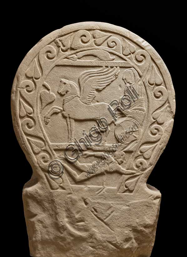 Museo Civico Archeologico di Bologna: stele funeraria etrusca decorata su entrambi i lati e nello spessore, lato A. Proveniente dal sepolcreto dei Giardini Margherita.inv. DUCATI_012Una donna dalla stirpe eroica. La stele Ducati 12, o di Rakvi Satlnei, è stata ritrovata presso la necropoli dei Giardini Margherita, collocata a ovest della città antica, ed è datata attorno al 400 a.C.  Questo lato è occupato dal tema del passaggio di status, cioè la morte, reso attraverso l’espediente del viaggio su carro della defunta. Sotto è raffigurato un personaggio maschile nudo che si getta su una spada, riconosciuto come l’eroe omerico Aiace Telamonio. Sulla stele è presente un’iscrizione che, in alcuni punti, è di difficile lettura. Si riesce a distinguere il prenome, Rakvi, e il gentilizio, Satlnei, della donna. È importante sottolineare che si tratterebbe dell’unico caso attestato in tutta Bologna. La seconda parte è molto dibattuta dalla critica ma sembra collegarsi alla raffigurazione di Aiace Telamonio. Infatti, le lettere leggibili sembrano comporre le parole aivas telmuns. Attraverso le informazioni grammaticali in nostro possesso, si è pensato che venga evocato un rapporto genealogico della defunta con l’eroe.