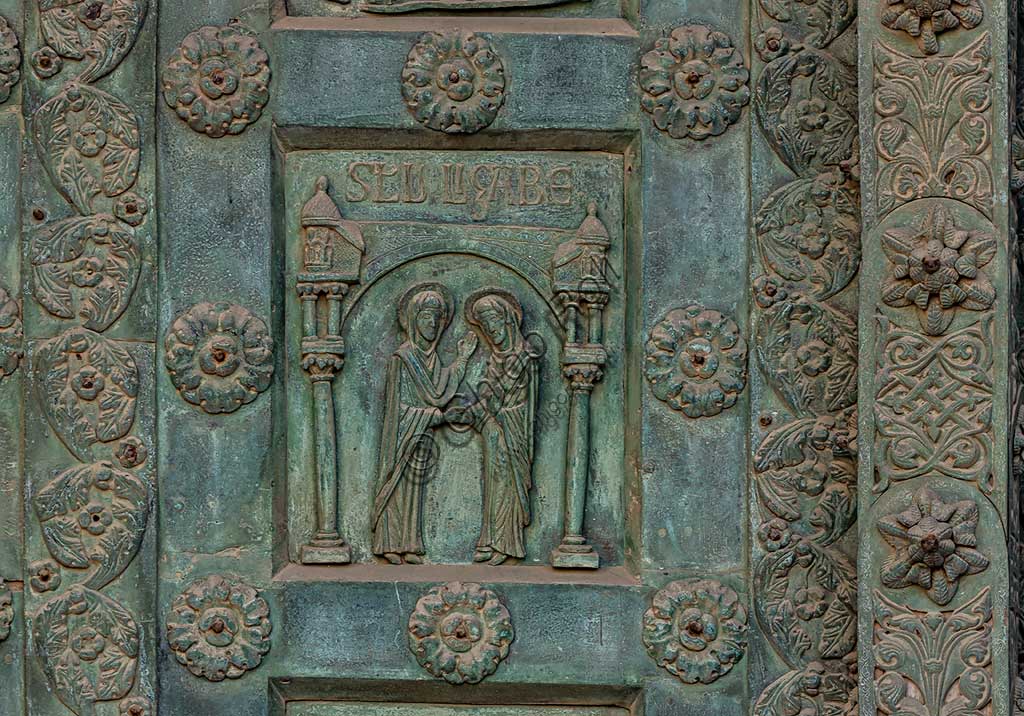 Duomo di Monreale, Porta di Bonanno Pisano (1185-6): formella bronzea con "Visitazione" (scena dal Nuovo Testamento).La porta è firmata: "Bonanno civis pisanus". Iconograficamente nei cinque ordini inferiori vi sono scene del Vecchio Testamento, sette dedicate alla storia di Adamo ed Eva, e altre sei ad altrettante coppie di profeti mentre nei cinque superiori scene del Nuovo, al sommo Cristo e Maria nella gloria del Paradiso.